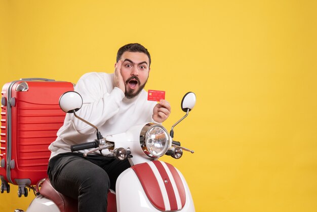 Concepto de viaje con joven viajero sorprendido sentado en motocicleta con maleta sosteniendo tarjeta bancaria en amarillo