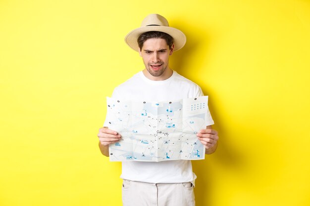 Concepto de viajes, vacaciones y turismo. Hombre mirando confundido en el mapa durante el viaje, no puedo entender, parado sobre fondo amarillo.