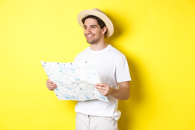 Concepto de viajes, vacaciones y turismo. Turista de chico guapo haciendo turismo, sosteniendo el mapa y sonriendo, de pie sobre fondo amarillo.