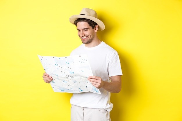 Concepto de viajes, vacaciones y turismo. Turista feliz sonriente mirando el mapa con visitas turísticas, de pie contra el fondo amarillo.