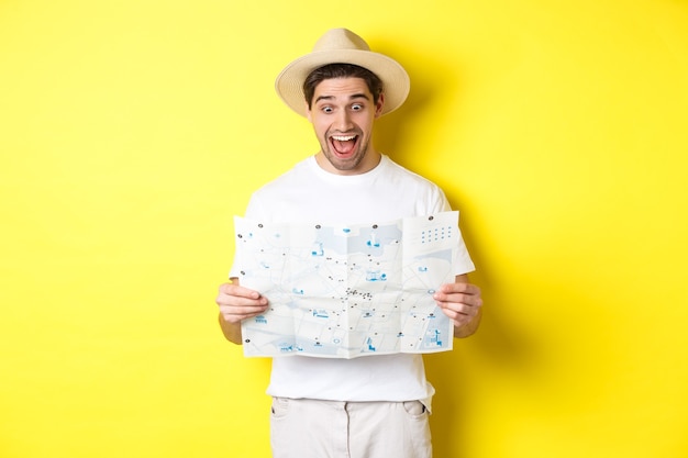 Concepto de viajes, vacaciones y turismo. Turista hombre mirando feliz en el mapa con suspiros, explorar la ciudad, de pie sobre fondo amarillo.