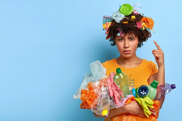 Concepto de voluntariado y protección del medio ambiente. Mujer afro disgustada apunta a la cabeza con desechos plásticos