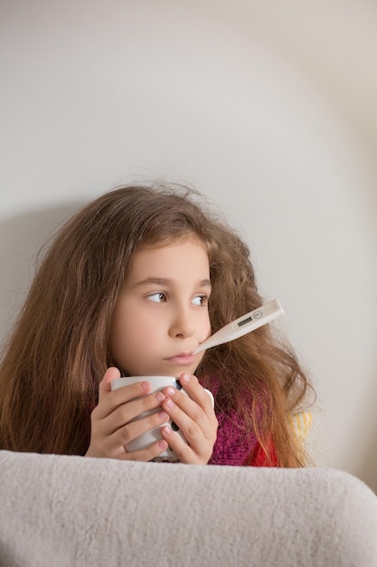 Conceptos de fiebre, resfriado y gripe Retrato de niña triste midiendo la temperatura del cuerpo y sosteniendo una taza de té frente a ella en casa