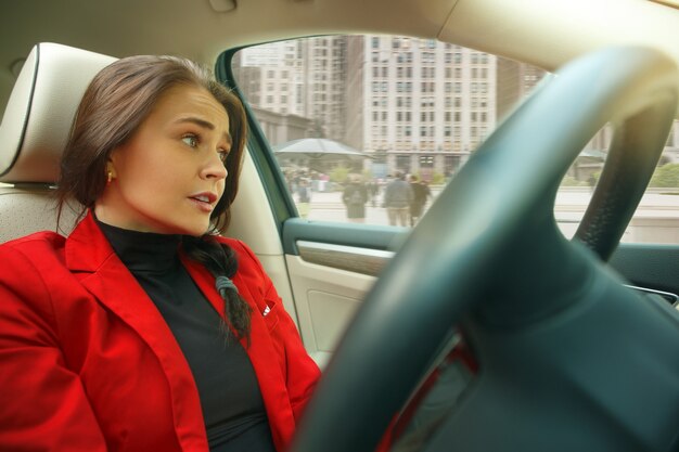 Conducir por la ciudad. Mujer atractiva joven conduciendo un coche. Joven modelo bastante caucásico en elegante chaqueta roja con estilo sentado en el interior del vehículo moderno. Concepto de empresaria.