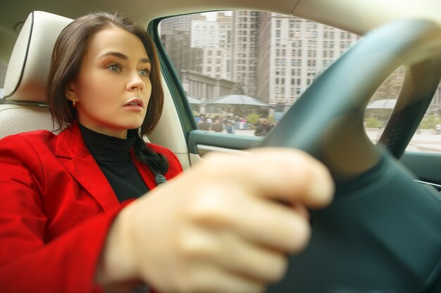 Conducir por la ciudad. Mujer atractiva joven conduciendo un coche. Joven modelo bastante caucásico en elegante chaqueta roja con estilo sentado en el interior del vehículo moderno