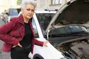 Foto gratuita conductor femenino maduro de pelo gris atractivo elegante de pie cerca de su coche blanco roto con el capó abierto