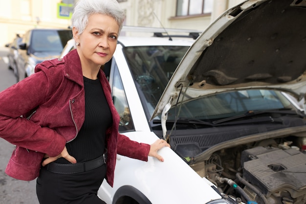 Conductor femenino maduro de pelo gris atractivo elegante de pie cerca de su coche blanco roto con el capó abierto