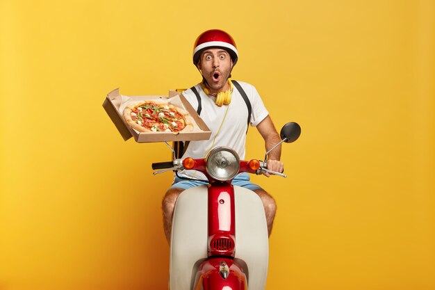 Conductor masculino guapo estupefacto en scooter con casco rojo entregando pizza