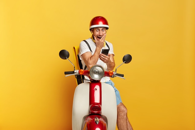 Conductor masculino guapo horrorizado en scooter con casco rojo