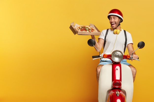 Conductor masculino guapo responsable en scooter con casco rojo entregando pizza