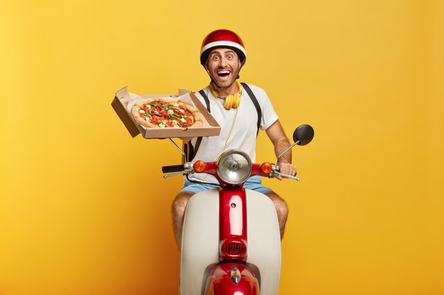 Conductor masculino guapo trabajador feliz en scooter con casco rojo entregando pizza