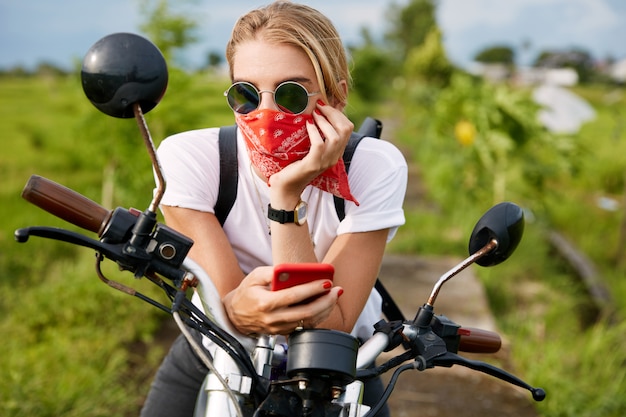 Conductora de moda vestida de manera informal, lee el blog de motociclistas en el teléfono móvil, se sienta en la motocicleta, refresca el aire fresco al aire libre, mira pensativamente a la distancia. Personas, estilo de vida y tecnología