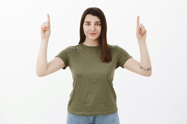 Confiada y ambiciosa, atractiva chica optimista con cabello castaño y tatuaje, sonriendo segura de sí misma y asertiva mientras levanta la mano y apunta hacia arriba pidiendo que eche un vistazo al espacio de la copia sobre una pared gris