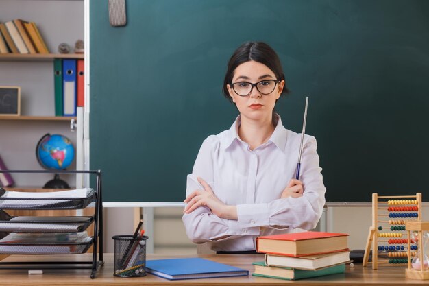 confiada cruzando las manos joven maestra con gafas sosteniendo un puntero sentado en el escritorio con herramientas escolares en el aula