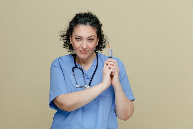 Confiada doctora de mediana edad con uniforme y estetoscopio alrededor del cuello sosteniendo una jeringa con aguja con ambas manos haciendo un gesto de pistola mirando la cámara aislada en un fondo verde oliva