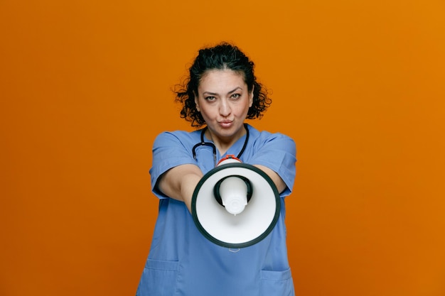 Confiada doctora de mediana edad usando uniforme y estetoscopio alrededor de su cuello mirando la cámara estirando el altavoz hacia la cámara aislada en el fondo naranja