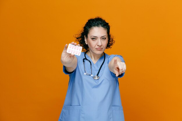 Foto gratuita confiada doctora de mediana edad usando uniforme y estetoscopio alrededor de su cuello mirando a la cámara estirando un paquete de cápsulas hacia la cámara apuntando a la cámara aislada en el fondo naranja