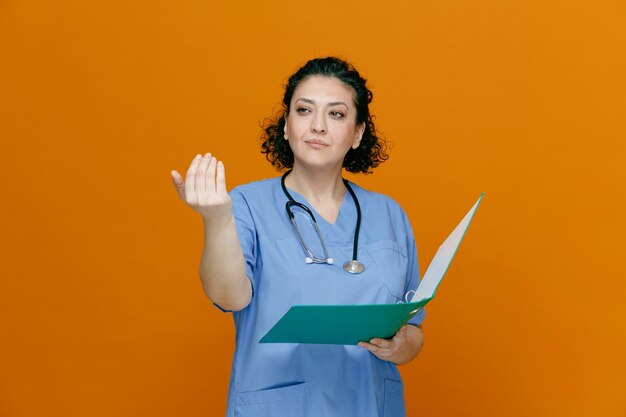 Confiada doctora de mediana edad usando uniforme y estetoscopio alrededor de su cuello sosteniendo una carpeta mirando al costado mostrando ven aquí gesto aislado en fondo naranja