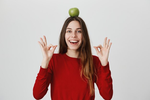 Confiada y emocionada, linda mujer sonriente mostrando un signo bien y sostenga la manzana verde en la cabeza