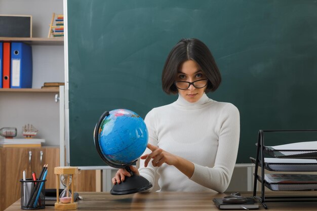 confiada mirando a la cámara joven maestra con gafas sosteniendo y apuntando al globo terráqueo sentada en el escritorio con herramientas escolares en el aula