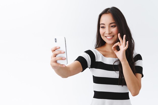 Confiada y saliente mujer bastante asiática morena en camiseta casual muestra un buen signo de aprobación en la pantalla móvil sosteniendo un teléfono inteligente tomando una tasa de selfie algo a través de una videollamada de fondo blanco