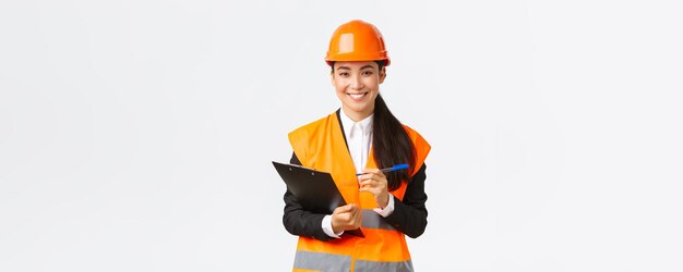 Confiada y sonriente ingeniera de construcción asiática mujer industrial en casco de seguridad visita el área del edificio para inspección escribiendo notas en el portapapeles y mirando satisfecho fondo blanco