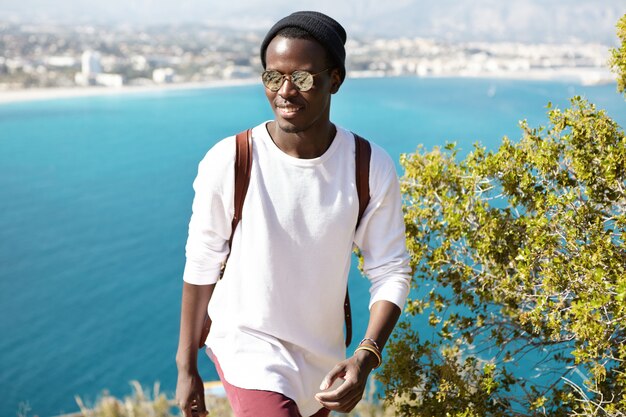 Confiado alegre joven estudiante europeo negro con gafas de moda y sombrero de senderismo en la montaña sobre el mar azul, mostrando a sus amigos bellezas y puntos de referencia de su ciudad natal en la costa