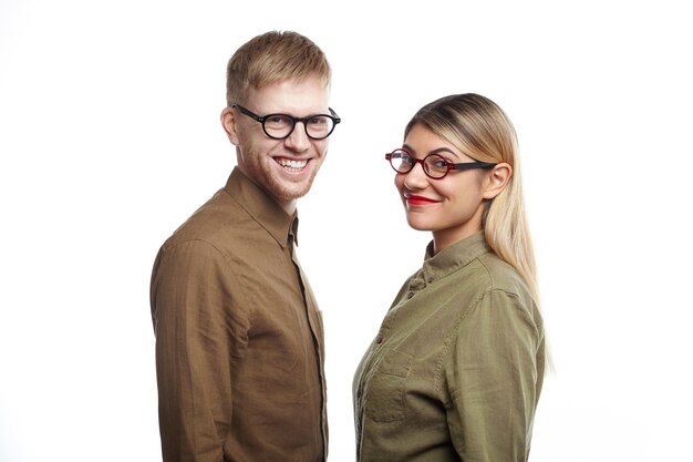 Confiado equipo exitoso de dos jóvenes socios comerciales ambiciosos masculinos y femeninos posando en la pared blanca y mirando con sonrisas alegres, felices con los buenos resultados de su proyecto común