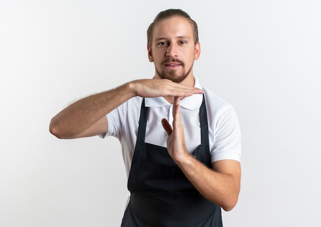 Confiado joven barbero guapo vistiendo uniforme haciendo gesto de tiempo de espera aislado en blanco con espacio de copia