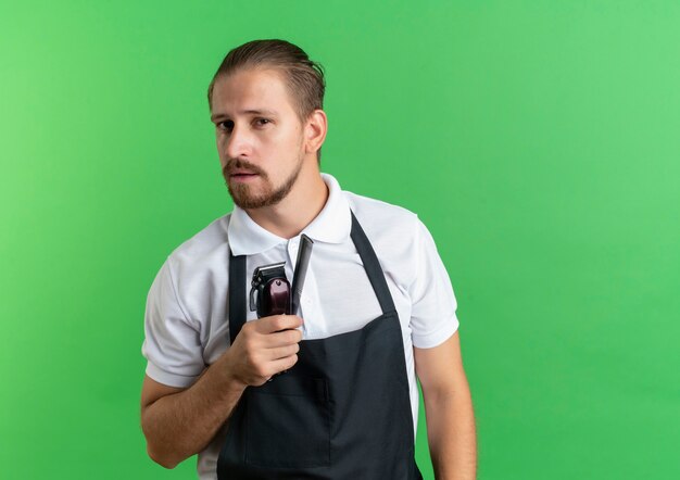 Confiado joven barbero guapo vistiendo uniforme con peine y cortapelos aislado en verde con espacio de copia