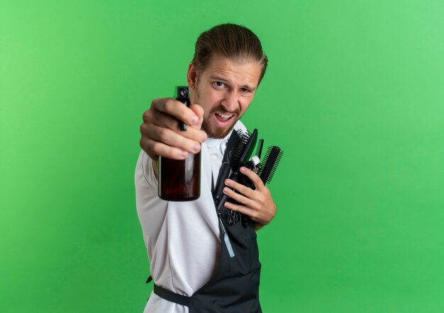 Confiado joven barbero guapo vistiendo uniforme de pie en la vista de perfil sosteniendo peines, cortapelos y estirando la botella de spray hacia la cámara aislada en verde con espacio de copia