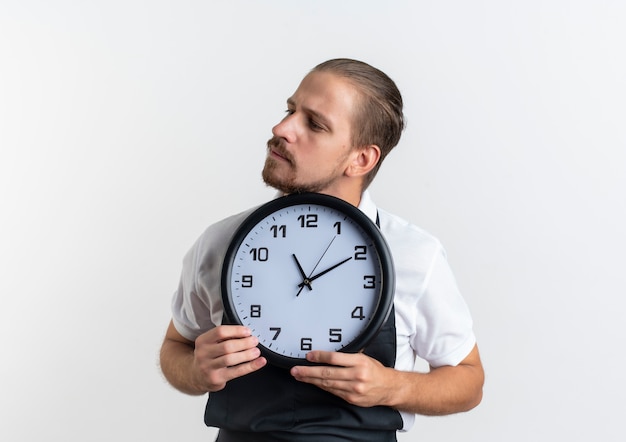 Confiado joven barbero guapo vistiendo uniforme sosteniendo el reloj y mirando al lado aislado en blanco con espacio de copia