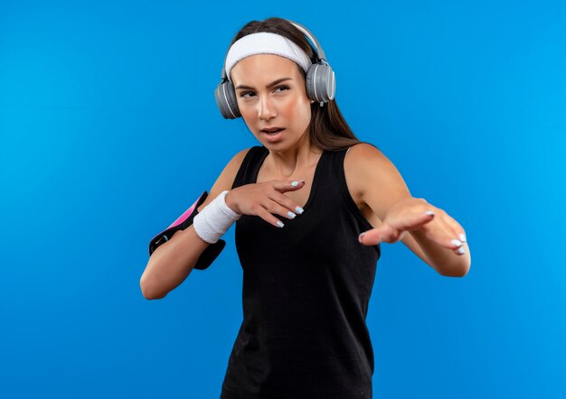 Confiado joven bastante deportivo vistiendo diadema y muñequera y auriculares con brazalete de teléfono estirando las manos mirando al lado aislado en la pared azul