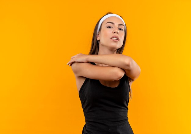 Foto gratuita confiado joven bastante deportivo vistiendo diadema y muñequera con los brazos cruzados en la pared naranja