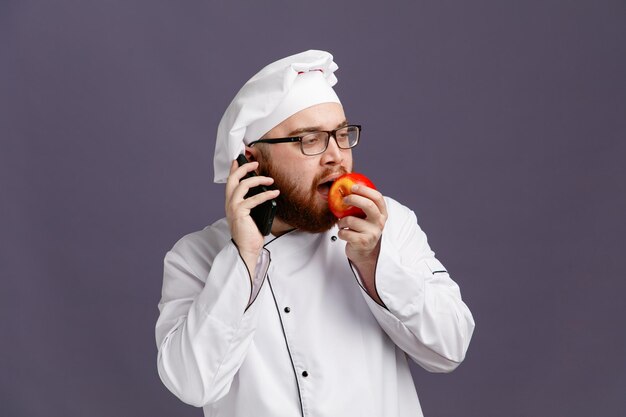 Confiado joven chef con anteojos uniformes y gorra mirando al costado comiendo manzana mientras habla por teléfono aislado en un fondo morado