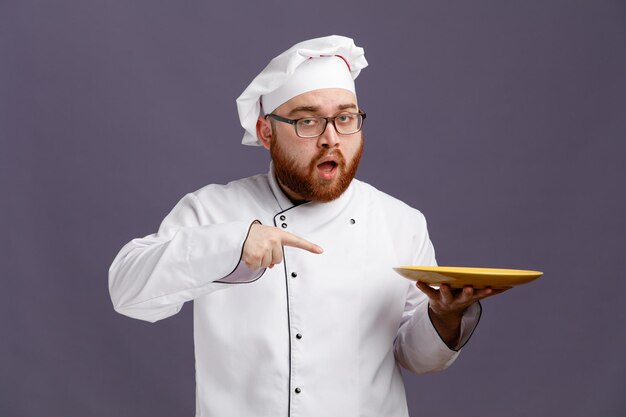 Confiado joven chef con anteojos uniformes y gorra sosteniendo y apuntando a un plato vacío mirando a la cámara aislada en un fondo morado