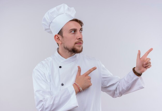 Un confiado joven chef barbudo hombre vestido con uniforme de cocina blanco y sombrero apuntando hacia arriba con los dedos índices mientras mira de lado en una pared blanca