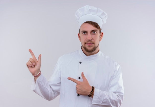 Un confiado joven chef barbudo hombre vestido con uniforme de cocina blanco y sombrero apuntando hacia arriba con los dedos índices mientras mira en una pared blanca