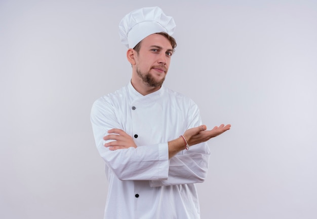 Un confiado joven chef barbudo hombre vestido con uniforme de cocina blanco y sombrero levantando la mano y tomados de la mano cruzados mientras mira en una pared blanca