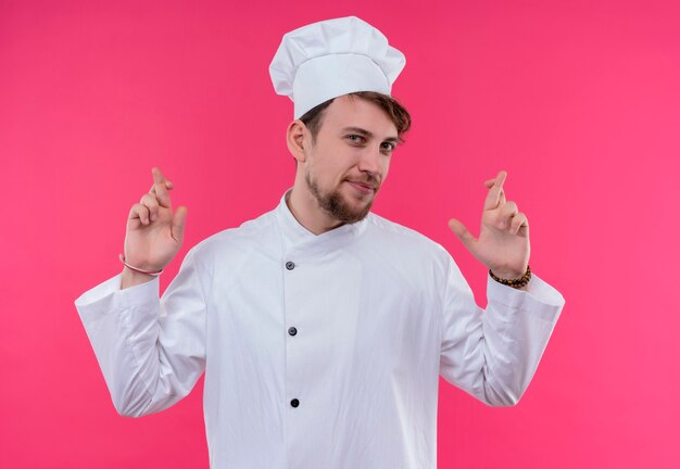 Un confiado joven chef barbudo en uniforme blanco mostrando los dedos cruzados mientras mira en una pared rosa