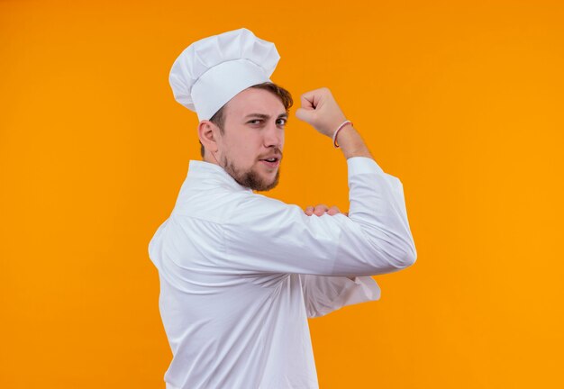 Foto gratuita un confiado joven chef barbudo con uniforme blanco mostrando sus músculos en una pared naranja