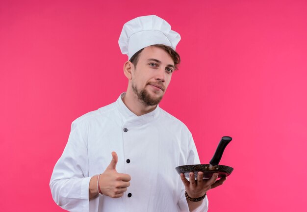 Un confiado joven chef barbudo con uniforme blanco que muestra los pulgares hacia arriba con una sartén en la otra mano mientras mira una pared rosa