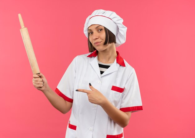 Confiado joven cocinera en uniforme de chef sosteniendo y apuntando al rodillo aislado en rosa con espacio de copia