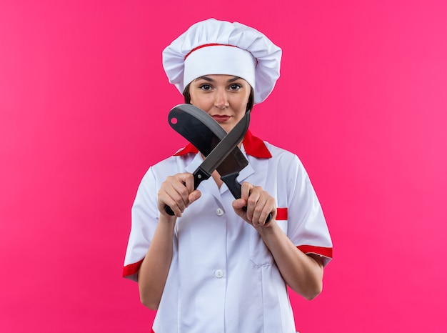 Confiado joven cocinera vistiendo uniforme de chef afilar cuchillo aislado sobre fondo de color rosa