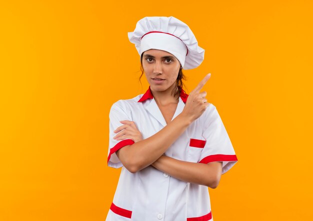 Confiado joven cocinera vistiendo uniforme de chef apunta al lado de la pared amarilla aislada con espacio de copia