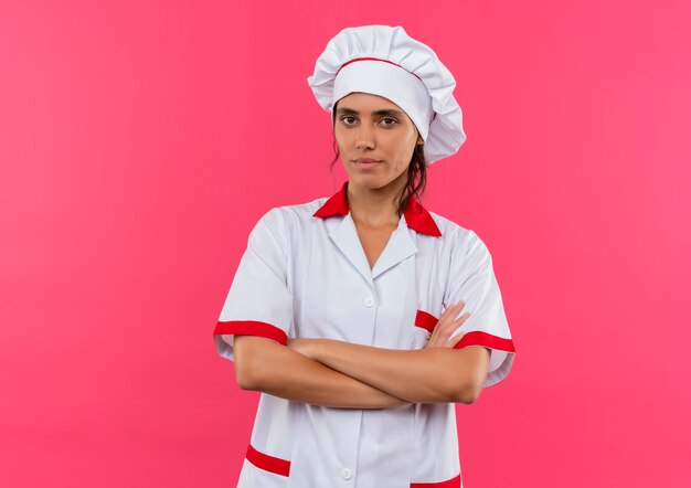 Confiado joven cocinera vistiendo uniforme de chef cruzando las manos en la pared rosa aislada con espacio de copia