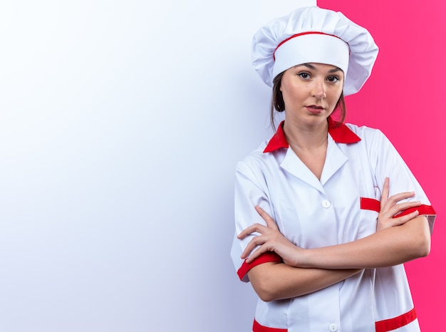 Confiado joven cocinera vistiendo uniforme de chef de pie delante de la pared blanca cruzando la mano aislada sobre fondo rosa con espacio de copia