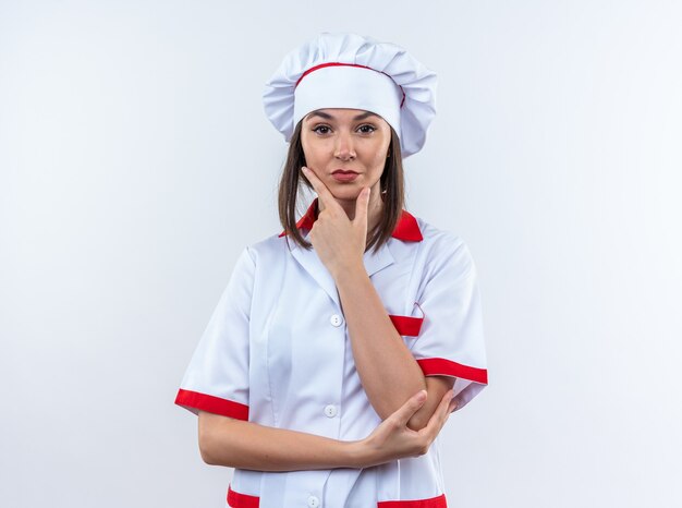 Confiado joven cocinera vistiendo uniforme de chef poniendo la mano en el mentón aislado en la pared blanca