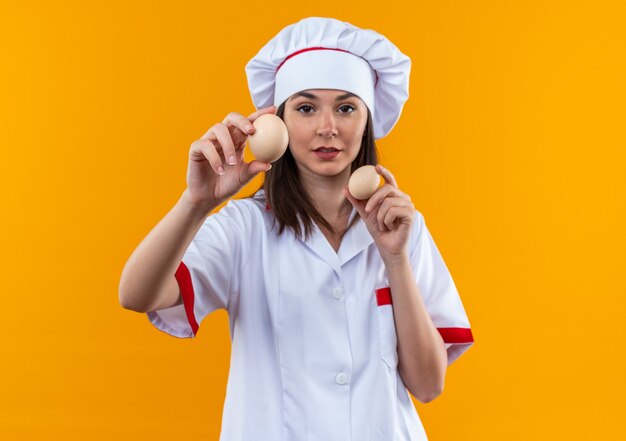 Confiado joven cocinera vistiendo uniforme de chef sosteniendo huevos en cámara aislada sobre fondo naranja