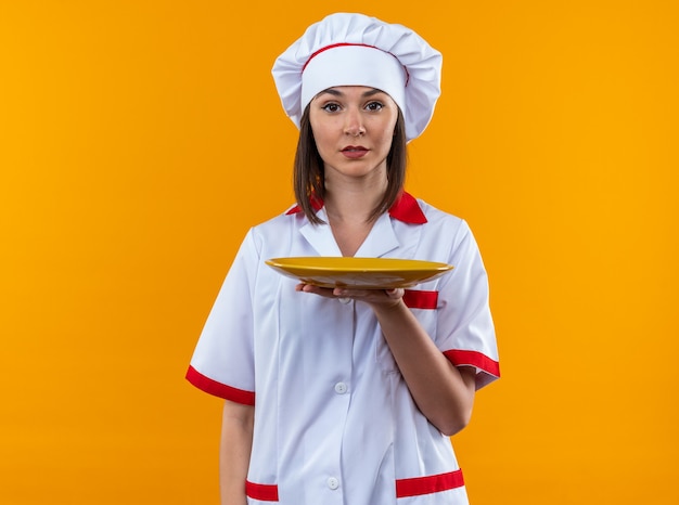Foto gratuita confiado joven cocinera vistiendo uniforme de chef sosteniendo la placa aislada sobre fondo naranja
